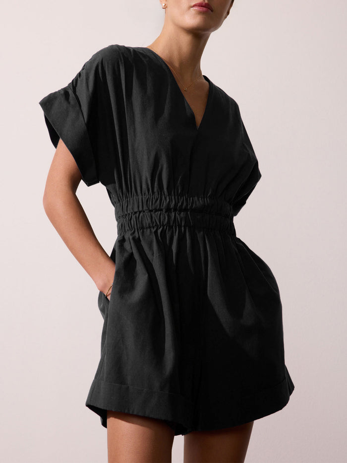 Front view of a woman wearing a black V-neck romper with a gathered elastic waist, against a beige studio background.