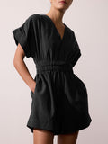 Front view of a woman wearing a black V-neck romper with a gathered elastic waist, against a beige studio background.