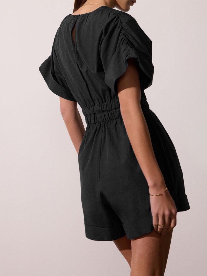 Woman wearing a black puff-sleeve romper with a gathered elastic waist, shown from a rear three-quarter angle against a beige studio background.