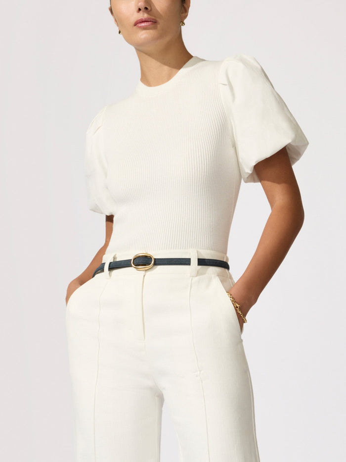 Woman wearing a white puff-sleeve knit top with white trousers and a navy belt with a gold buckle