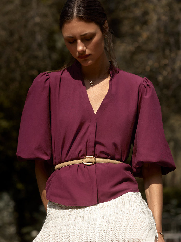 Woman wearing a plum puff-sleeve Marla blouse with a tan belt over a cream knit skirt