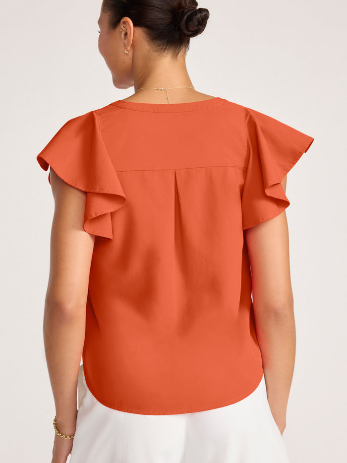 Woman wearing a rust orange ruffle-sleeve button-front blouse with white pants, back view