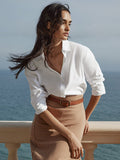 Woman with windblown hair wearing a white button-up shirt and camel knit skirt with a brown belt by the ocean