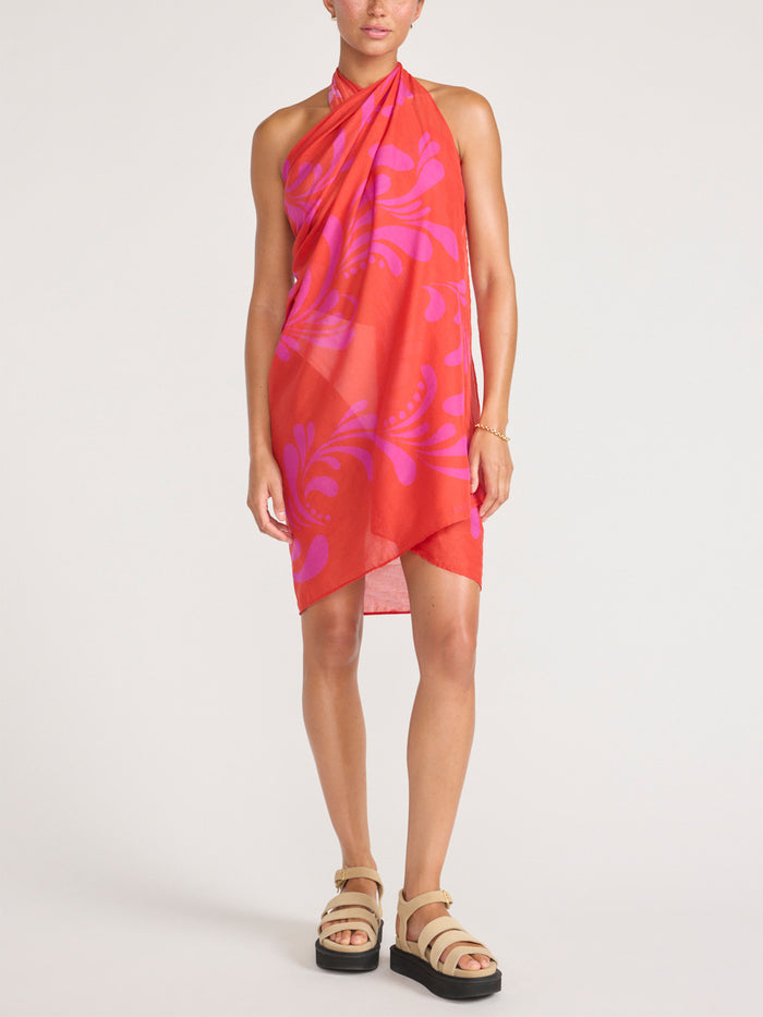 Front view of a model wearing the orange and pink tropical-print pareo styled as a halter wrap dress, with platform sandals.