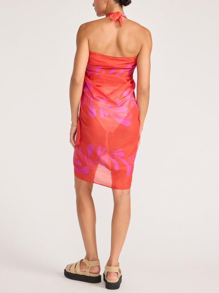 Back view of a model wearing the orange and pink tropical-print pareo styled as a halter wrap dress, with platform sandals.