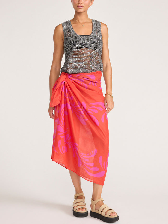 Front view of a model wearing an orange and pink tropical-print pareo wrap skirt with a knit tank top and platform sandals.