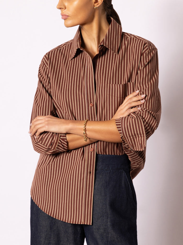 Woman wearing a brown and beige vertically striped button-up shirt with dark blue jeans, against a light gray studio background.