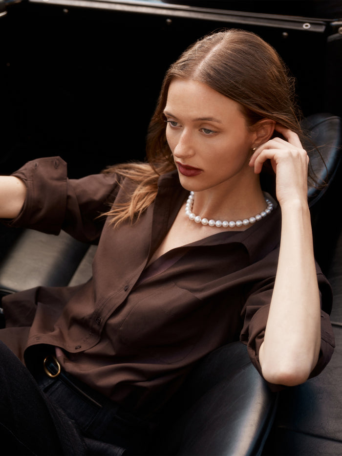 Model in a chocolate brown silk button-up shirt and pearl necklace, seated in a car