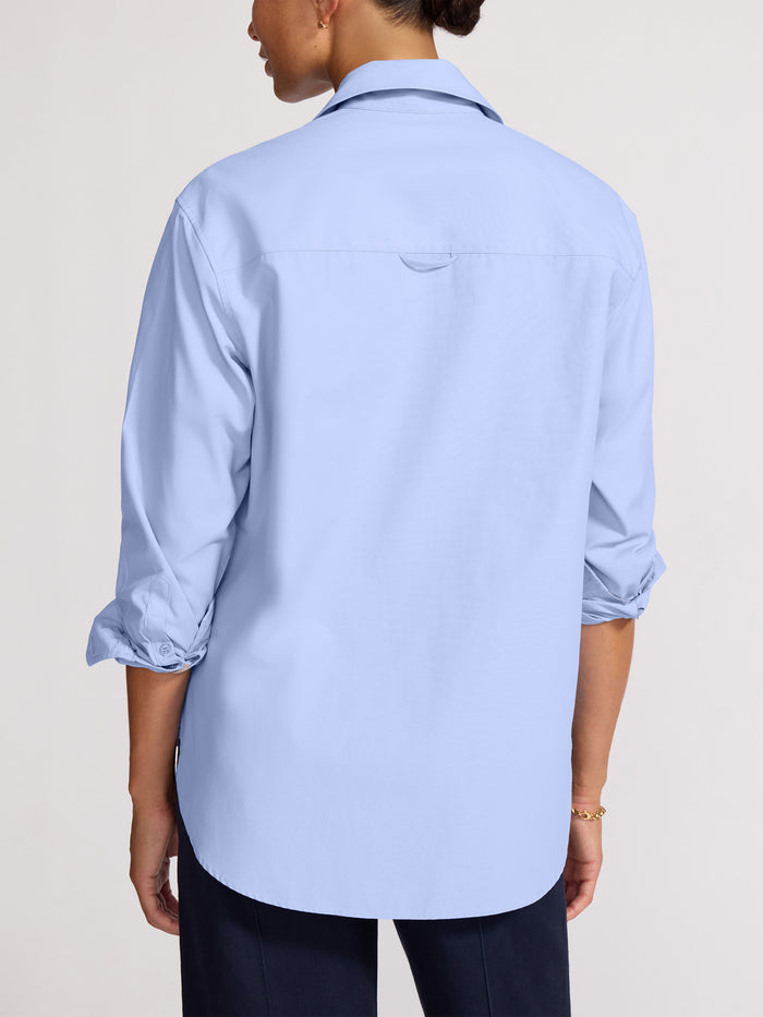 Back view of a woman wearing a light blue button-front shirt with rolled-up sleeves over navy trousers