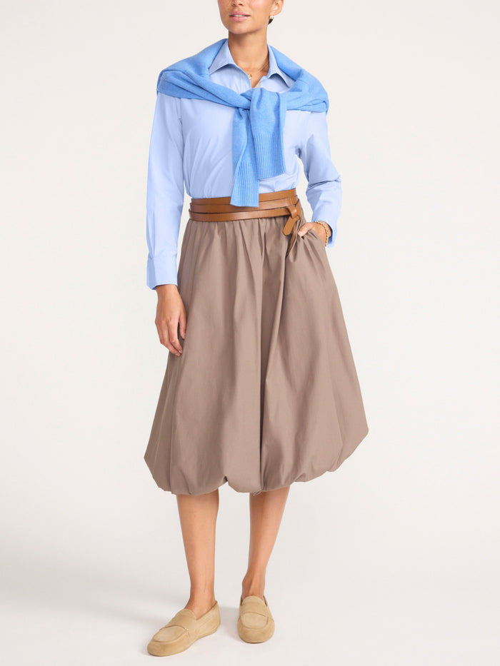 Full-length front view of a woman wearing a light blue button-front shirt with a sweater tied at the shoulders, a belted taupe balloon midi skirt, and tan loafers