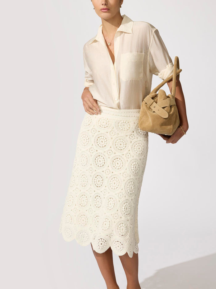 Woman wearing an ivory blouse with a white crochet midi skirt with scalloped hem, holding a tan suede bag