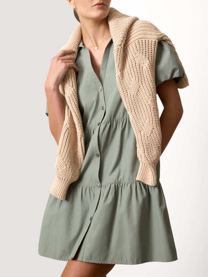 Woman wearing a sage green tiered shirt dress with a beige knit sweater draped over her shoulders