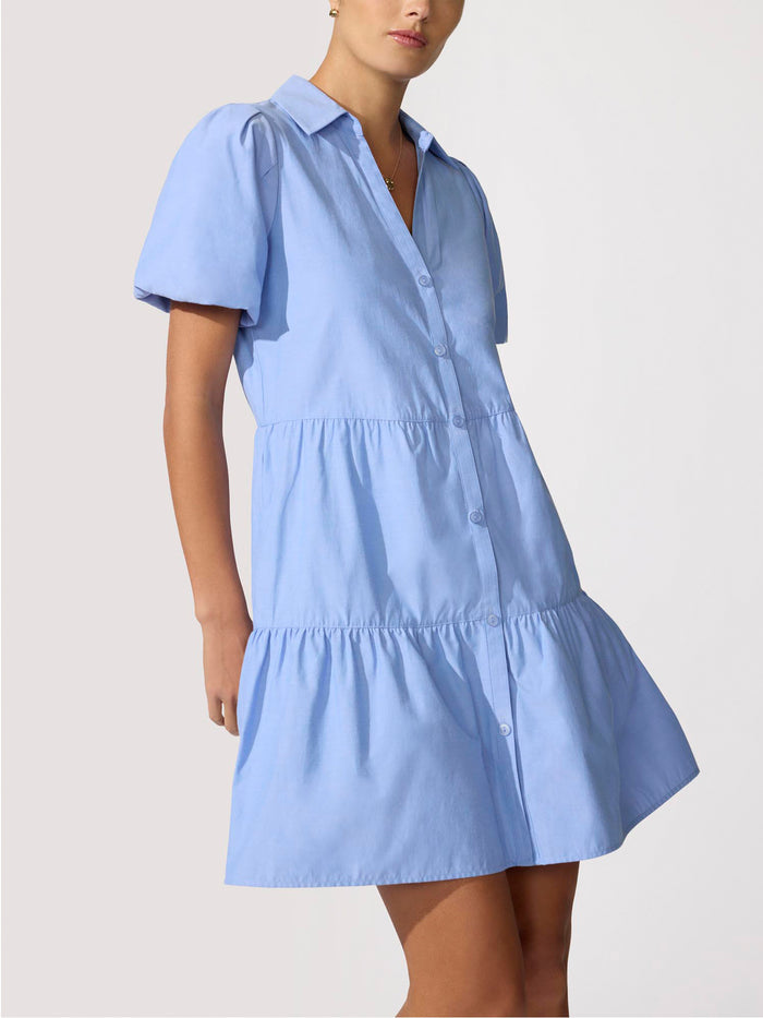 Woman wearing a periwinkle blue tiered ruffle mini shirtdress with puff sleeves and front pockets.