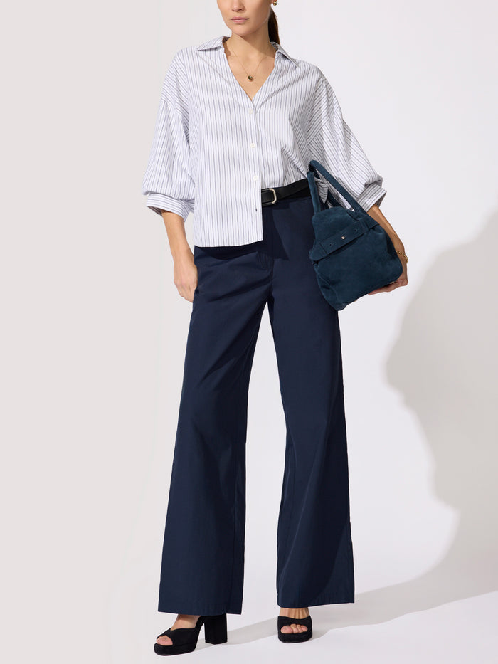 Woman styled in an oversized white striped button-down shirt tucked into wide-leg navy trousers, carrying a teal suede tote bag.