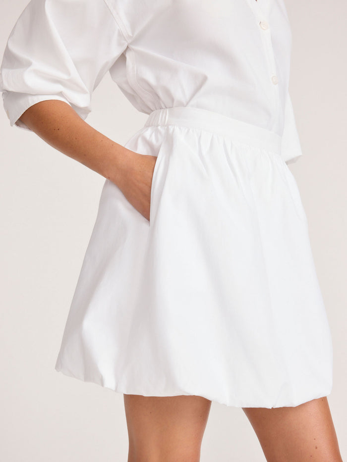 Close-up side view of a model wearing a white bubble mini skirt with a white blouson top, hand in pocket