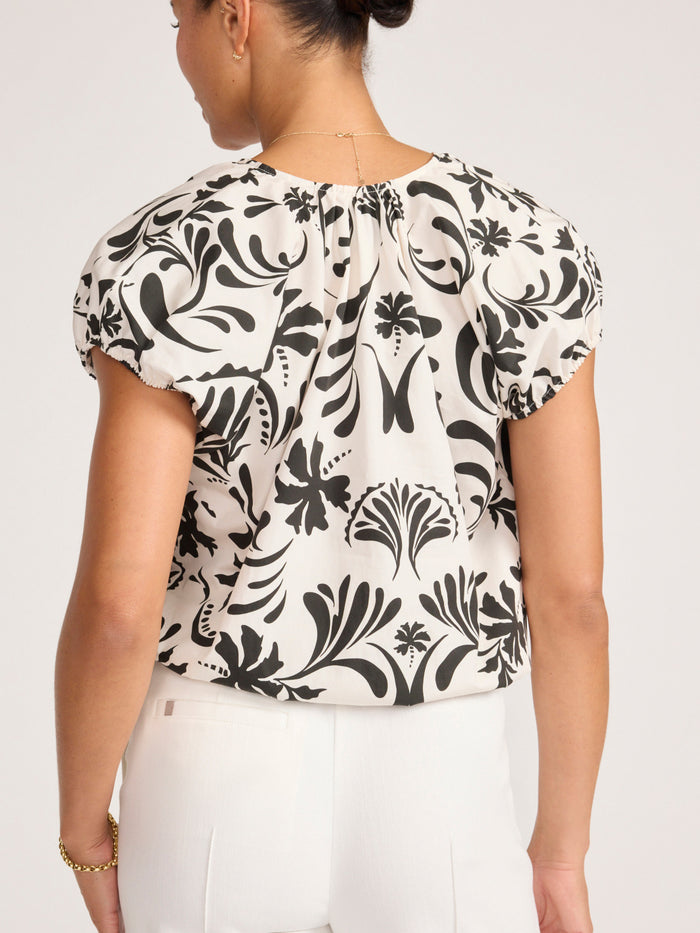 Woman wearing a black and white floral print blouse with balloon sleeves and white trousers, back view