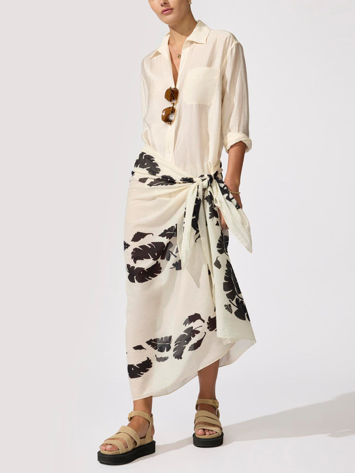 Woman wearing a cream button-down shirt dress with a black palm-leaf print sarong-tied skirt