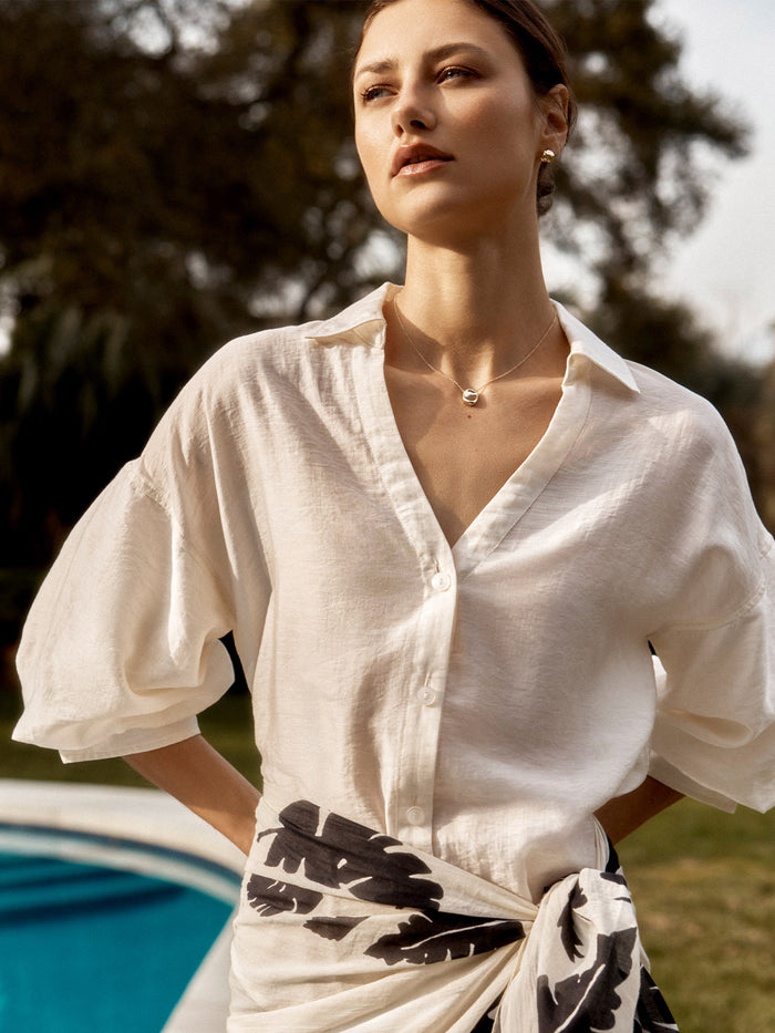 Close-up of a woman wearing a cream button-down shirt with a black palm-leaf print sarong skirt by a pool