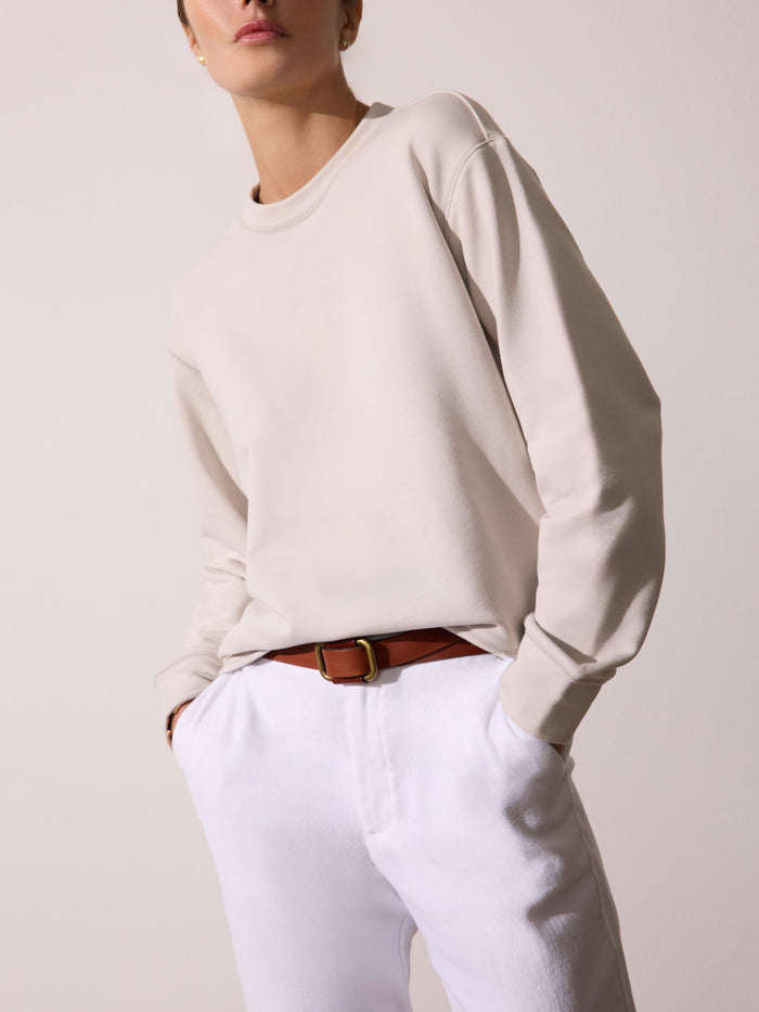 Model wearing a cream crewneck sweatshirt, white trousers, and a brown leather belt with a gold buckle, against a light beige background.