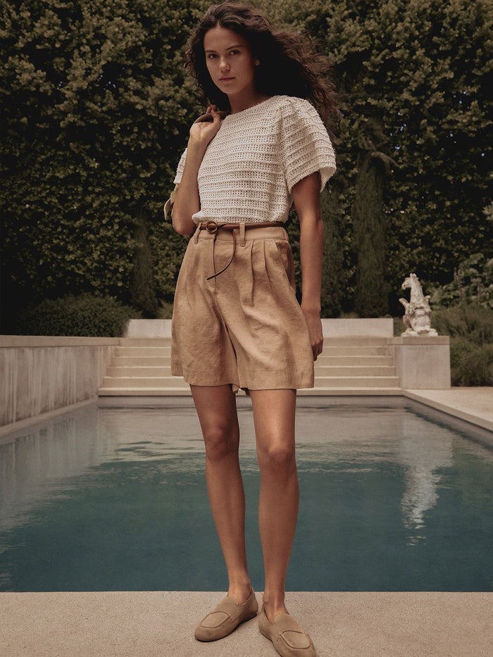 Woman wearing a white crochet knit top with tan pleated Maris shorts, standing poolside