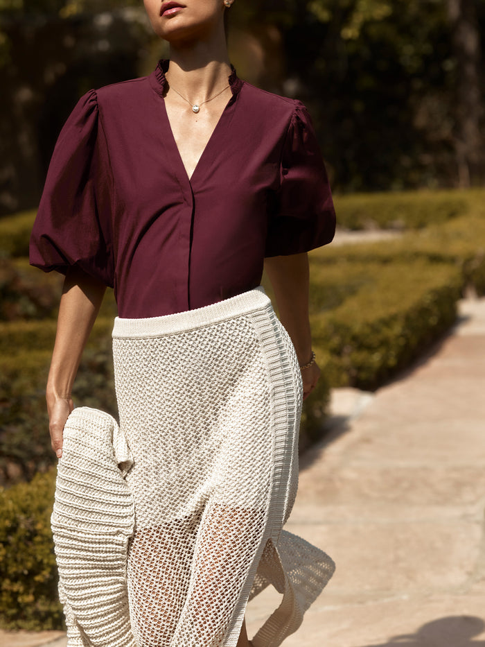 Woman wearing a burgundy puff-sleeve blouse with a cream crochet knit maxi skirt, walking through a garden