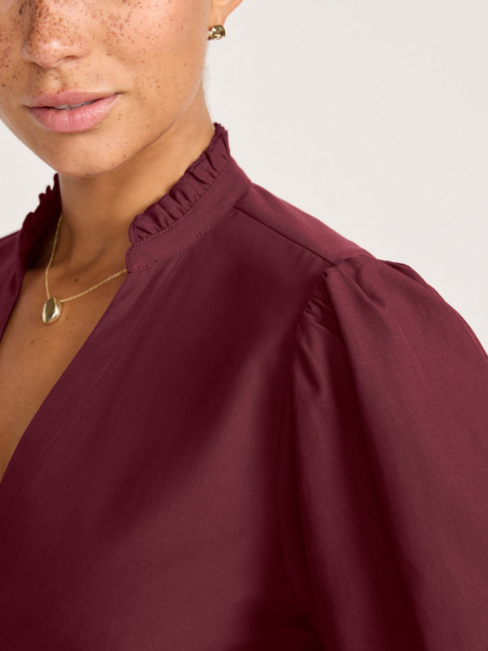 Close-up of a woman wearing a burgundy puff-sleeve blouse with a ruffled collar and a gold pendant necklace