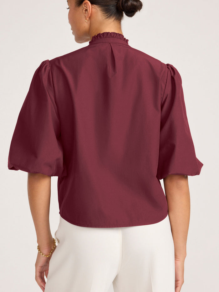 Back view of a woman wearing a burgundy puff-sleeve blouse with a ruffled collar and cream trousers