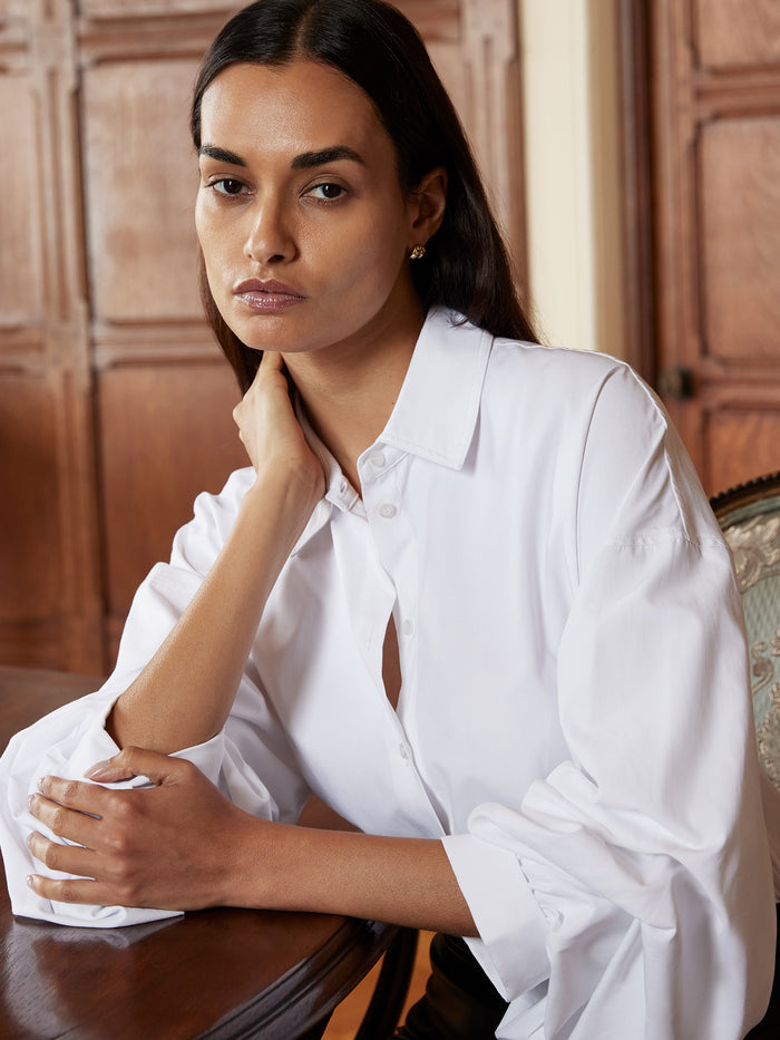 Woman with long dark hair wearing an oversized white button-up shirt with voluminous sleeves, seated in front of a wood-paneled background.