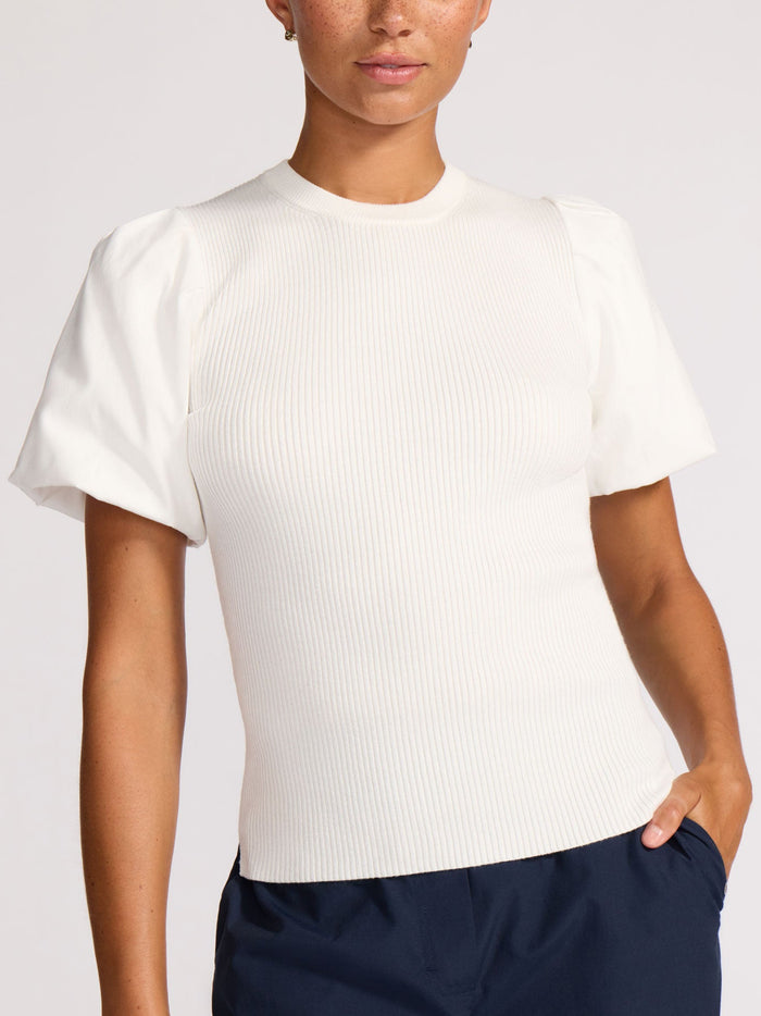 Woman with hand in pocket wearing a white ribbed knit top with puff sleeves and navy trousers