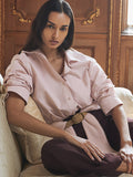Model seated wearing a blush pink button-up shirt tied at the waist with a tan belt and burgundy trousers