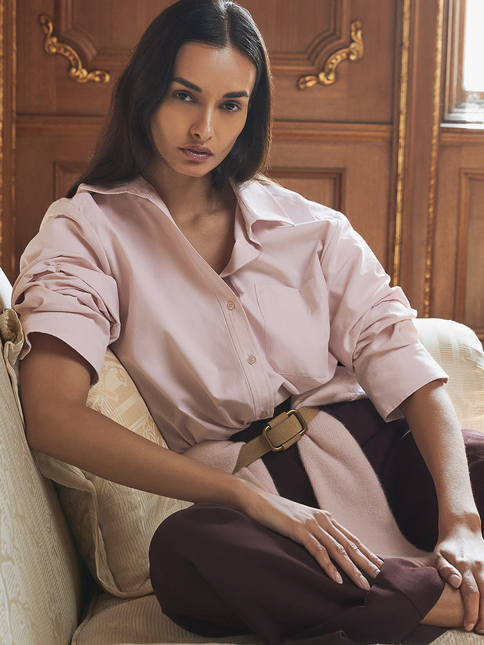 Model seated wearing a blush pink button-up shirt tied at the waist with a tan belt and burgundy trousers