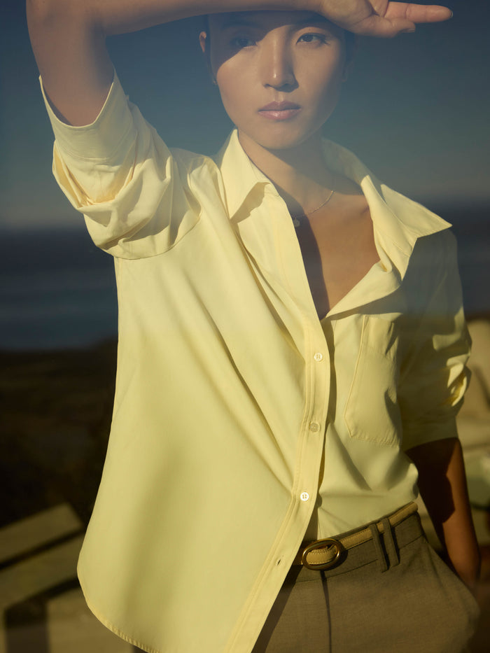 Woman shielding her eyes wearing a pale yellow button-down shirt and olive trousers by the coast
