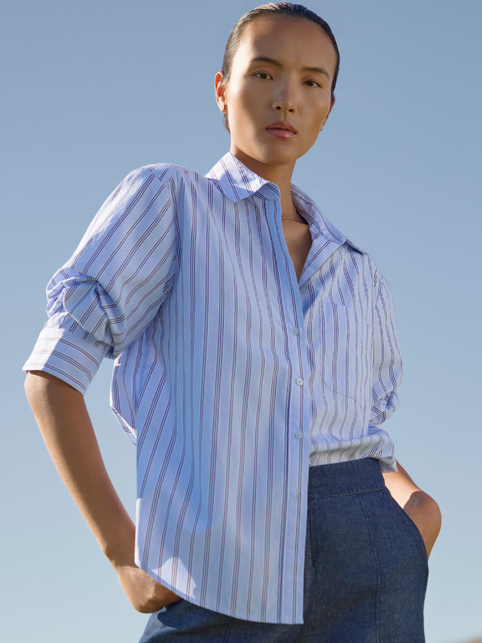 Close-up of a woman with hands in her pockets wearing a blue and white striped button-down shirt and dark denim pants