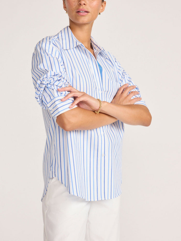 Model with arms crossed wearing a blue and white striped button-up shirt and white trousers
