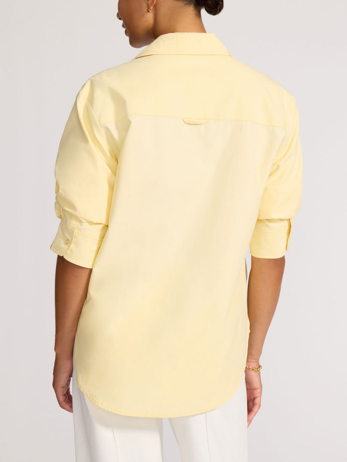 Back view of a model wearing a pale yellow button-down shirt with rolled sleeves tucked into white trousers