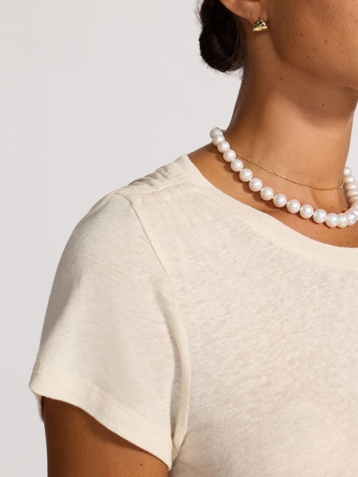 Close-up of a cream linen crew-neck t-shirt worn with a pearl necklace and gold stud earrings.
