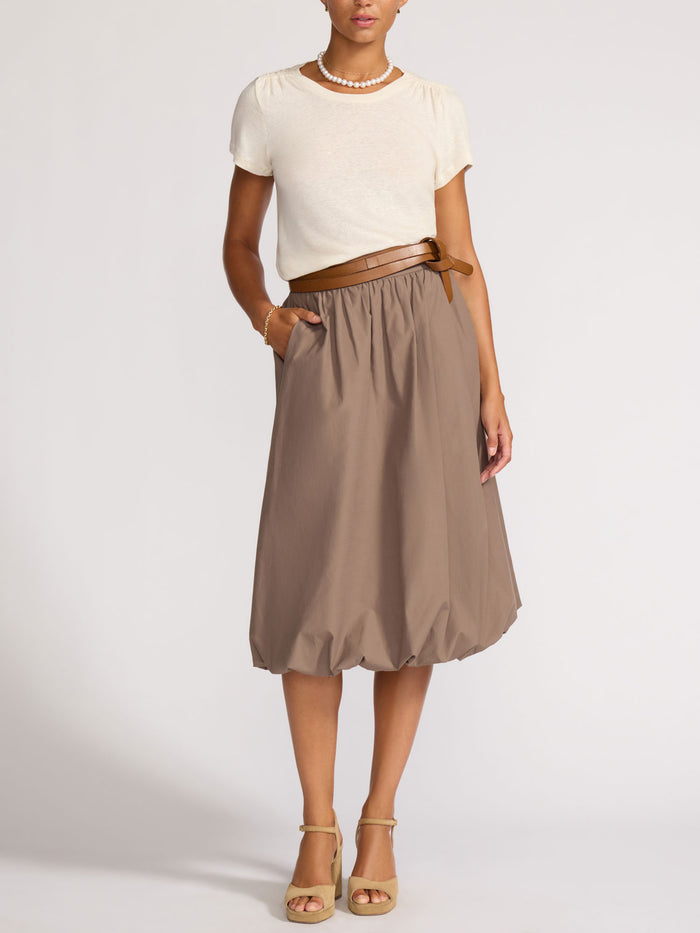 Woman wearing a cream linen t-shirt tucked into a taupe midi skirt with a brown leather belt and tan sandals.