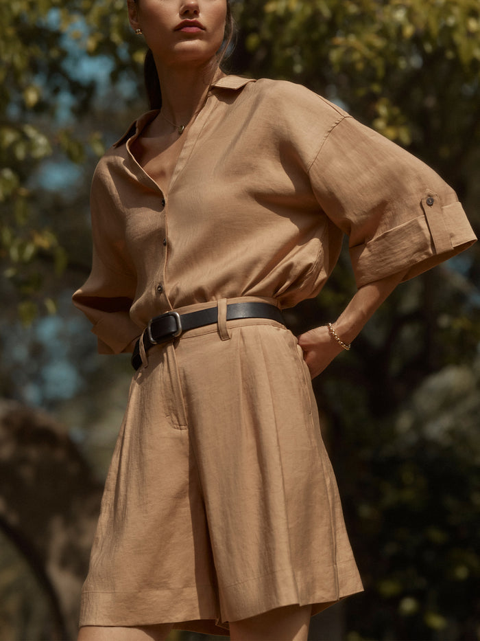 Woman wearing a desert tan Ryder linen shirt tucked into pleated Maris shorts with a black belt