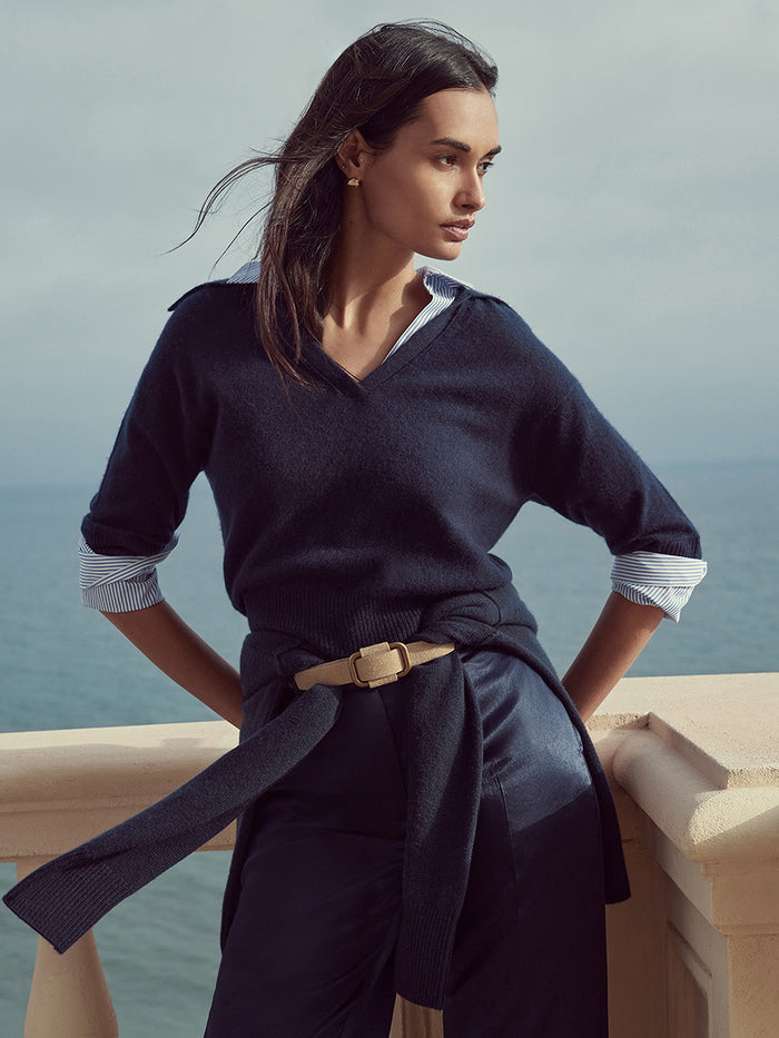 Woman wearing a navy V-neck sweater over a striped shirt with a tan belt, standing by the ocean